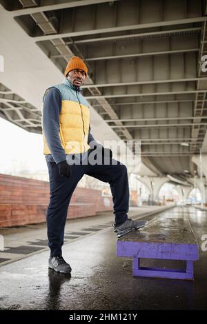 Junger ernsthafter blackman in warmem Trainingsanzug und ärmelloser Jacke, der den Fuß auf der Bank hält, um auf dem Sportplatz zu trainieren Stockfoto