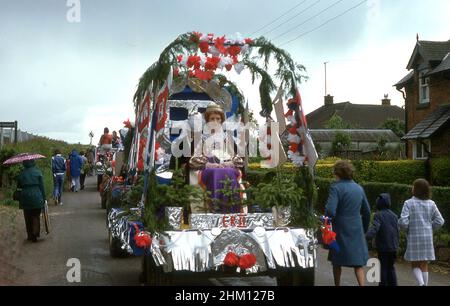 1977, historisch, auf einer engen englischen Dorfstraße in einer Parade von Festwagen, eine Frau in Kostümen als Königin sitzend auf einem dekorierten Parade-Festwagen mit rot-blauem Bund, zur Feier des silbernen Jubiläums von Königin Elizabeth II. Die Einheimischen gehen die Gasse hinauf zum Feld, wo die Feierlichkeiten stattfinden werden. Stockfoto