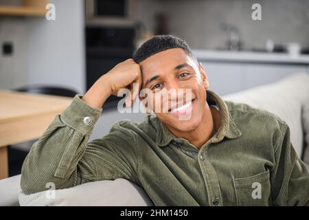 Porträt eines gutaussehenden jungen Mannes, der sich zu Hause im modernen Wohnzimmer ausruhen kann, während er lässig auf dem Sofa sitzt, die Kamera anschaut und sich entspannt fühlt. Lifestyle- und Freizeitkonzept Stockfoto