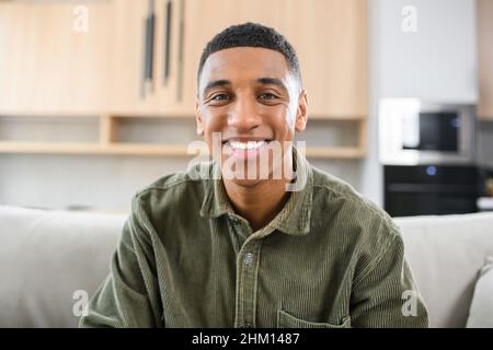 Porträt eines sorglosen jungen Mannes, der auf dem Sofa sitzt und die Kamera anschaut. Heiterer Mann in legerem Hemd in entspannter Atmosphäre, der Freizeit zu Hause verbringt Stockfoto