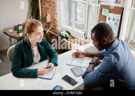 Zwei junge, selbstbewusste interkulturelle Kollegen mit Geschäftsdokumenten, die bei einem Arbeitstreffen über Arbeitsplan oder Daten diskutieren Stockfoto