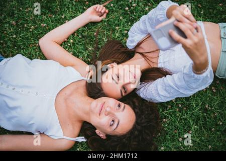 Zwei Freunde machen ein Selfie-Foto, während sie sich in einem Park auf dem Gras niederlegen. Stockfoto