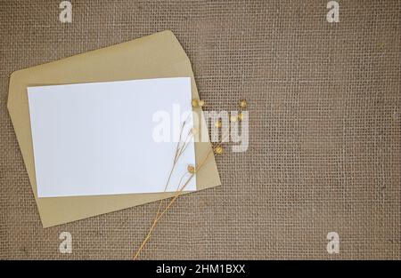 Modell mit Umschlag und leerer Karte. Ein Umschlag aus Kraftpapier auf Sackleinen. Getrocknete Blüten (trockener Flachs mit Samenkapseln) neben dem Umschlag. Stockfoto