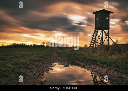 Jagdturm, im Sonnenuntergang mit sich bewegenden Wolken und Reflexen und einer Wasserpfütze Stockfoto