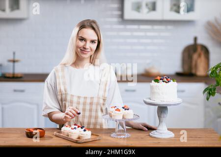 Konditorei Konditorin junge kaukasische Frau in beigefarbener Schürze mit Kuchen auf Küchentisch. Kuchen Cupcakes und süßes Dessert Stockfoto