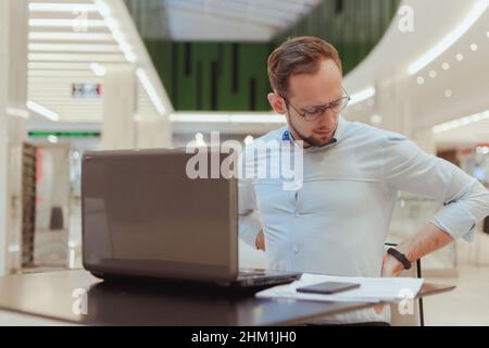 Der Mann hält sich am Rücken fest, leidet unter Schmerzen im unteren Rückenbereich und arbeitet am Computer. Sitzende Büroangestellte. Stockfoto