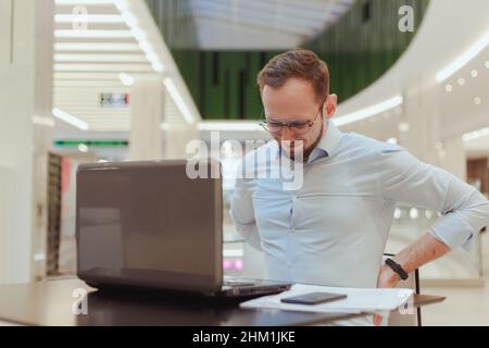 Der Mann hält sich am Rücken fest, leidet unter Schmerzen im unteren Rückenbereich und arbeitet am Computer. Sitzende Büroangestellte Stockfoto