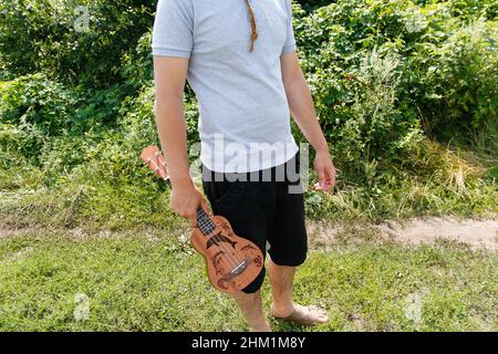 Musikhobby. Junger schöner Kaukasischer, der Ukulele draußen hält. Schöner Hipster Kerl spielt Ukulele Gitarre auf grünem Natur Hintergrund. Männlich in einem CO Stockfoto
