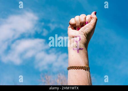 Faust einer älteren Frau mit lila lackierten Nägeln, mit dem Himmel im Hintergrund, mit dem weiblichen Symbol bemalt. Konzept des Frauentages, Empowerment, Stockfoto