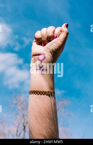 Faust einer älteren Frau mit lila lackierten Nägeln, mit dem Himmel im Hintergrund, mit dem weiblichen Symbol bemalt. Konzept des Frauentages, Empowerment, Stockfoto