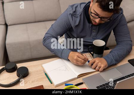 Kleiner Latino-Junge, der seine Notizen in ein Notizbuch auf dem Tisch neben seinem Laptop und seiner Kaffeetasse schreibt. Stockfoto