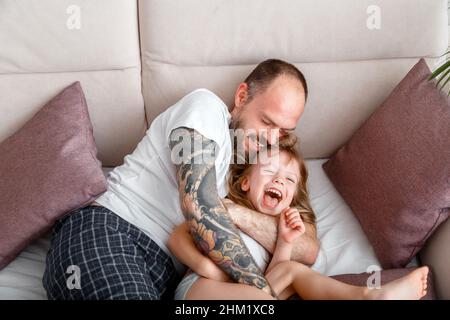 Vater spielen mit Kind Kind Mädchen verbringen Zeit zusammen. Kahlbärtiger Kaukasischer Mann mit Tattoos kitzelt seine zarte kleine Tochter. Mädchen lacht spielen Stockfoto