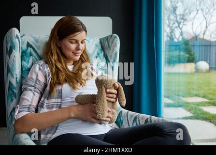 Charmante Schwangere, die ein Baby erwartet, sich auf einem Sessel in der Nähe des Fensters mit Blick auf den Garten entspannt und ein Plüsch-Kaninchenspielzeug auf ihrem schwanger Bauch hält, e Stockfoto