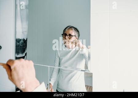 Die alte Frau wäscht ihre Zähne vor einem Spiegel mit einer Bambuszahnbürste Stockfoto