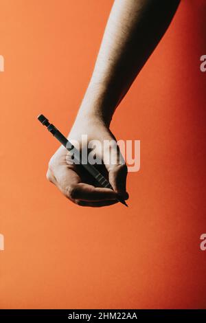 Eine junge Hand, die einen mechanischen Bleistift auf einem orangefarbenen Hintergrund mit tiefen Schatten hält Stockfoto