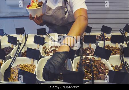 der verkäufer gießt Beläge auf einen weichen, gefrorenen Joghurt in einer weißen Take-Away-Tasse Stockfoto