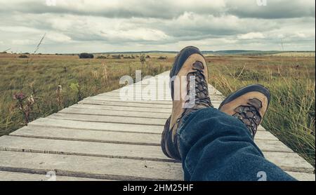 Wanderschuhe eines Wanderers, während Sie sich auf einer hölzernen Promenade ausruhen, aus Sicht. Stockfoto