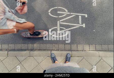 Ein junger Mann, der an einer Bordwand in der Stadt steht, und eine Frau fahren mit einem Mini Cruiser Board auf die Straßen. Sichtbild. Stockfoto