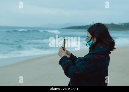 Frau, die während eines stürmischen Tages ein Selfie mit einer Grippemaske am Strand gemacht hat Stockfoto