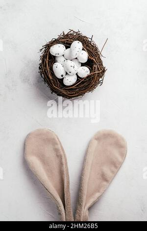 Osterzusammensetzung aus weißen Wachteleiern in braunem Nest auf weiß grauem Papierhintergrund und Ausschneiden von beigefarbenem Hasen-Kaninchen-Spielzeug flauschige Haarohren. Cele Stockfoto