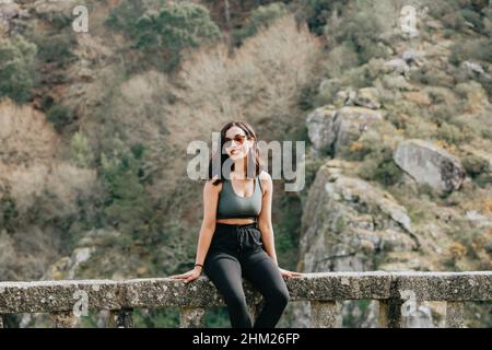 Junge arabische Frau in Sportkleidung lächelt mit Sonnenbrillen, während sie an einem Herbsttag von der Kamera in einen Wald blickt Stockfoto