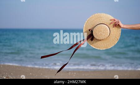 Nahaufnahme eines Strohhuts mit Bändern, die im Wind in einer weiblichen Hand vor dem Hintergrund von Meer, Meer, Strand, Kopierraum flattern. Konzept des Urlaubs, Stockfoto