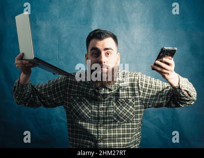 Junger Hipster-Mann mit Bart-Konzept, der mit einem Laptop und einem Telefon auf blauem Hintergrund arbeitete, betonte den Platz für die Kamerakopie Stockfoto
