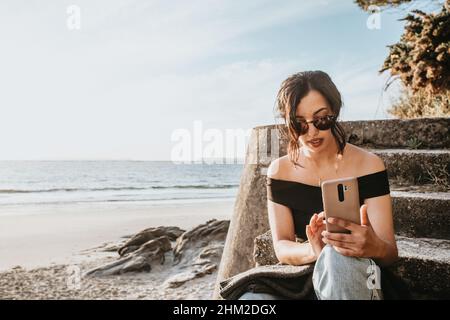 Junge Frau in Sommerkleidung und Sonnenbrille mit seinem Handy lächelnd mit Kopierplatz am Strand Stockfoto