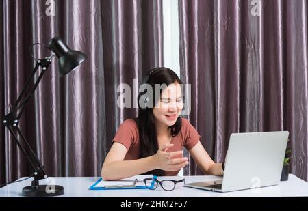 Arbeit von zu Hause, asiatische junge Geschäftsfrau lächeln mit Kopfhörern Videokonferenz oder facetime von Laptop-Computer sie heben die Hand zu erklären Stockfoto