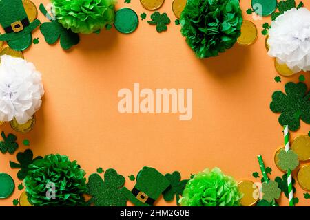 „Happy St. Patricks Day“-Kartendesign. Rahmen aus Kleeblättern, Goldmünzen, grünen Dekorationen auf orangefarbenem Hintergrund. Flach liegend, Draufsicht. Vinta Stockfoto