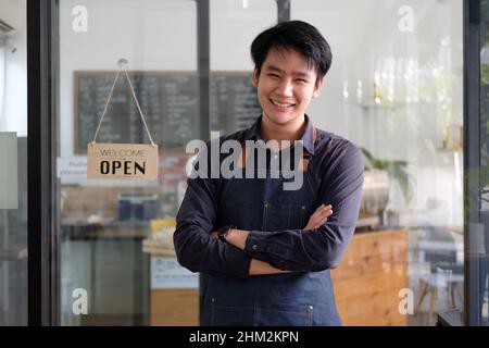 Fröhlicher junger, asiatischer Barista, der auf der Schürze ein Tablet hält und vor der Tür des Cafés mit offenem Schild steht. Unternehmer Startup SME Stockfoto