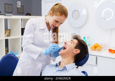 Ärztin in weißer Robe und Latexhandschuhen lächelnd und mit den Zähnen des fröhlichen Mannes, der in der modernen Zahnklinik arbeitet Stockfoto
