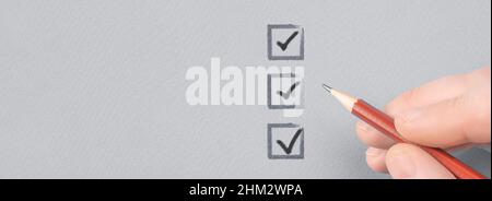 Hand mit Bleistift schreiben Häkchen auf Checkliste. Hand mit Bleistift und Kontrollkästchen auf grauem Papierhintergrund. Männliche Hand wählt drei von drei Optionen Stockfoto