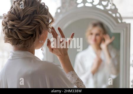 Hautpflege für Frauen. Kaukasische Frau berührt ihr Gesicht und sieht im Wohnzimmer in einem seidenweißen Bademantel in einem Sessel am Fenster im Spiegel aus. Genießt ihre Reflexion mit schönen Haaren und Make-up Stockfoto