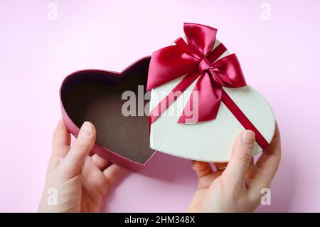 Die Hände der Frau öffnen die Geschenkbox auf rosa Hintergrund, Draufsicht. Leere Geschenk herzförmige Box große rote Schleife verziert. Flache Lay-Komposition zum Geburtstag, Stockfoto
