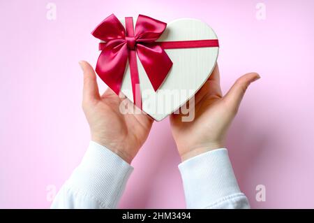 Womans Hände halten Geschenk oder Geschenk herzförmige Box dekoriert große rote Schleife auf rosa Tisch Draufsicht. Flache Lay-Komposition zum Geburtstag, Valentinstag Stockfoto