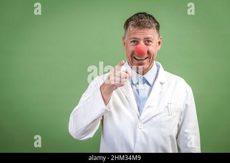 Wissenschaftler mit weißem Mantel hat rote Clownnase auf und steht vor grünem Hintergrund Stockfoto