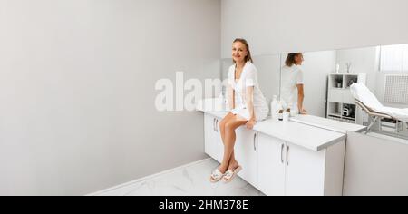 Eine junge lächelnde Kosmetologin sitzt auf einem Tisch in einem medizinischen Büro. Speicherplatz kopieren. Das Konzept der professionellen Kosmetologie. Stockfoto