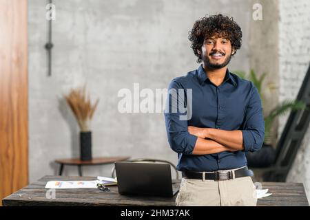 Lächelnder, lockiger Geschäftsmann aus dem Nahen Osten, der neben dem Schreibtisch steht und die Kamera anschaut. Junge positive männliche Studentin mit moderner Wohnung auf Hintergrund. Stolzer und erfolgreicher Inhaber von Kleinunternehmen mit gemischtem Rennen Stockfoto
