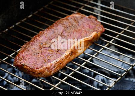 1 rohes Steak (Rumpsteak) von Rindfleisch liegt auf einem Edelstahl-Grillrost. Glühende Kohlen rösten die Delikatesse. Blick von schräg vorne. Stockfoto