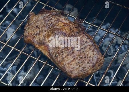 1 Steak (Rumpsteak) von Rindfleisch liegt auf einem Edelstahlgrill. Glühende Kohlen rösten die Delikatesse.leichte Rauchentwicklung. Blick von oben. Stockfoto