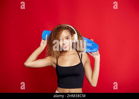 Fröhliche afroamerikanische Hipsterin mit lockigen Haaren mit Penny Board, die Musik in Kopfhörern hört und im Studio vor rotem Hintergrund auf die Kamera schaut Stockfoto