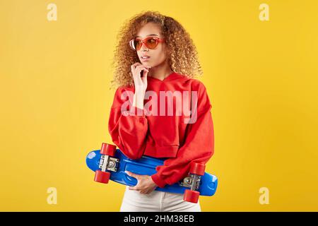 Stilvolles afroamerikanisches Hipster mit lockigen Haaren in Sonnenbrille und Penny Board, das vor gelbem Hintergrund steht und das Kinn berührt Stockfoto