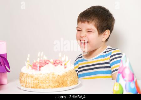 Ein kleiner Junge lacht, feiert seinen Geburtstag, sitzt mit Kerzen vor einem weißen Kuchen. Stockfoto