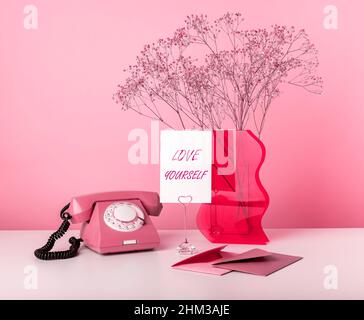 Karte mit Worten Liebe dich selbst. Retro-Telefon, getrocknete Blumen in Vase, Umschläge mit Grußkarten am Tisch auf rosa Hintergrund. Selbstachtung oder Valentinstag-Konzept. Hochwertige Fotos Stockfoto