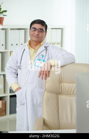 Porträt eines selbstbewussten lächelnden Chefarztes, der an seinem Stuhl im medizinischen Büro steht Stockfoto