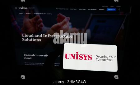 Person, die das Smartphone mit dem Logo des US-AMERIKANISCHEN IT-Unternehmens Unisys Corporation auf dem Bildschirm vor der Website hält. Konzentrieren Sie sich auf die Telefonanzeige. Stockfoto