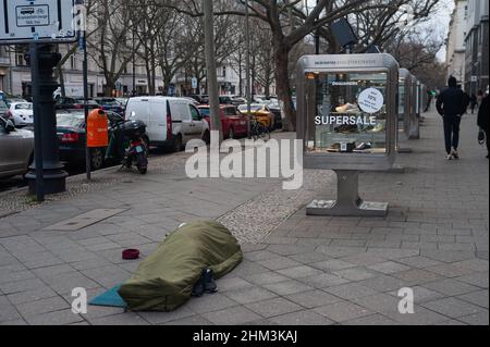 05.02.2022, Berlin, Deutschland, Europa - Obdachloser schläft in seinem Schlafsack auf dem Bürgersteig vor einem Modehaus am Kurfürstendamm. Stockfoto