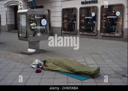 05.02.2022, Berlin, Deutschland, Europa - Obdachloser schläft in seinem Schlafsack auf dem Bürgersteig vor einem Modehaus am Kurfürstendamm. Stockfoto