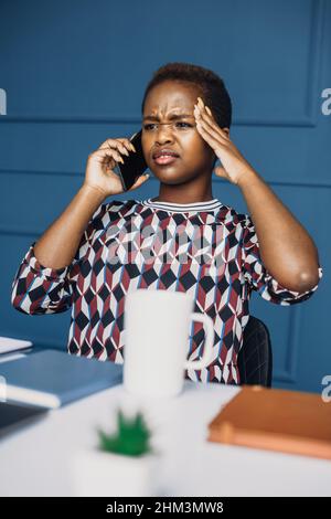 afroamerikanische Frau, die auf dem Smartphone sprach und auf dem Tisch saß, betonte bei seinem neuen Job. Porträt einer Geschäftsfrau. Sprechende Menschen. Stockfoto